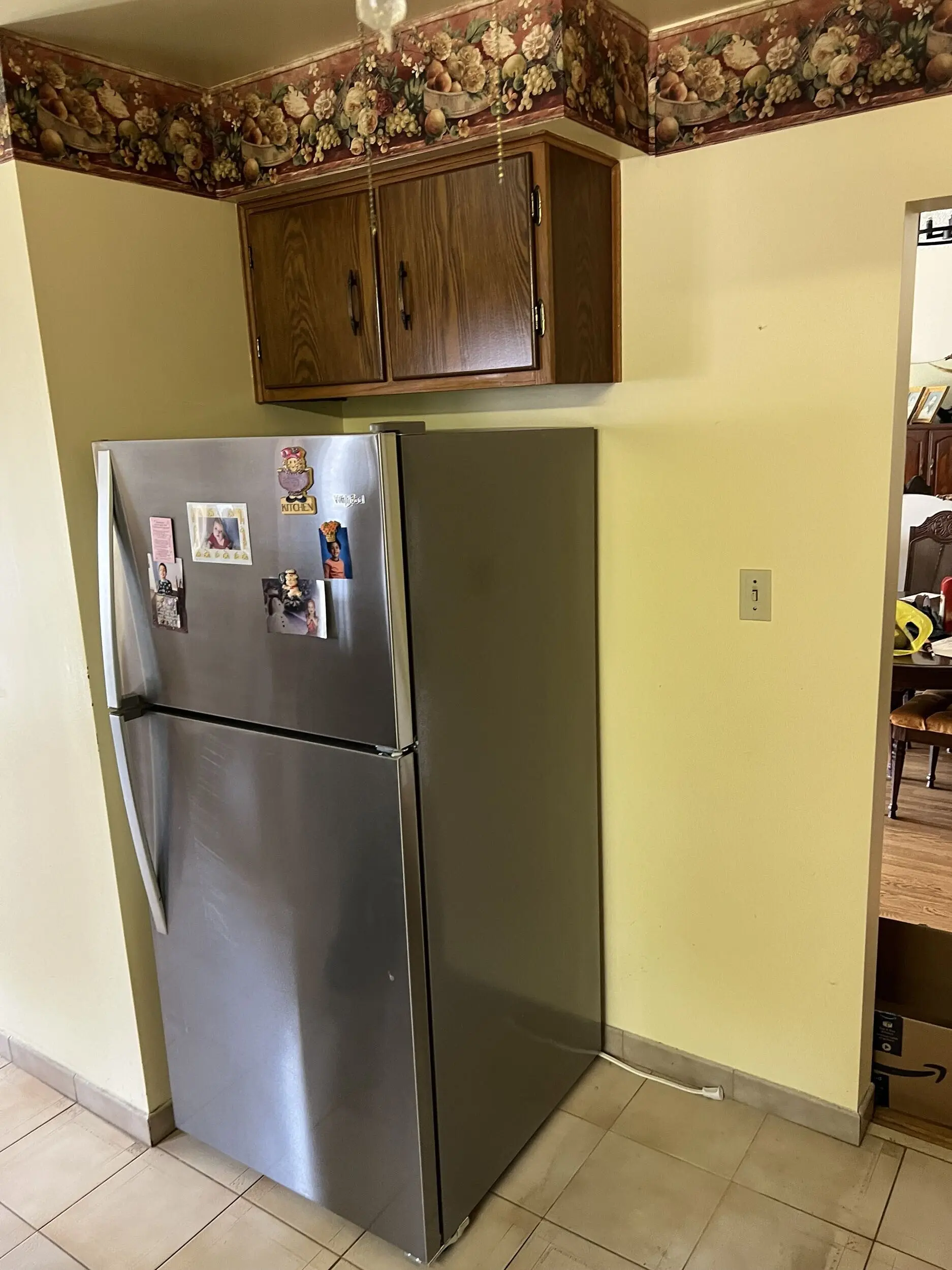 A before photo of a Stainless steel refrigerator with various magnets and photos on the door, positioned against a yellow wall in a kitchen corner, with wooden cabinets above and floral-patterned wallpaper border near the ceiling.