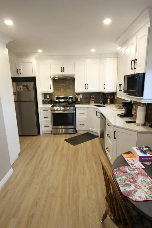 Modern kitchen with white cabinetry, stainless steel appliances including refrigerator, stove, microwave, and dishwasher, light wooden flooring, and a round dining table with a wooden chair, placemats, and some books on the side.