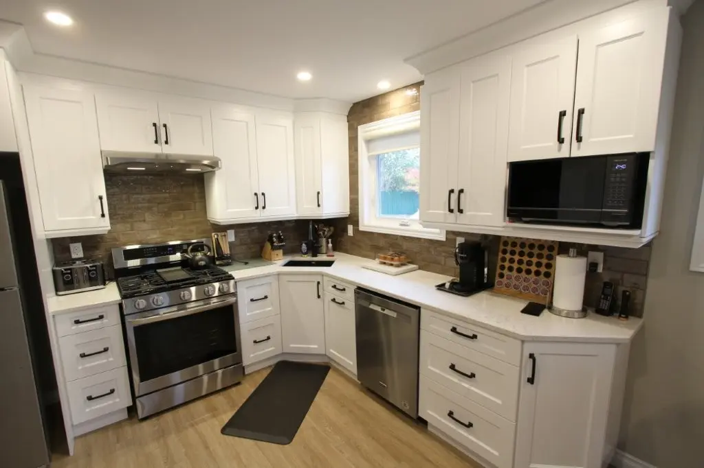 Modern kitchen with white cabinets, stainless steel appliances including a stove, dishwasher, and microwave, light wood flooring, a window above the counter, and a brick-patterned backsplash.