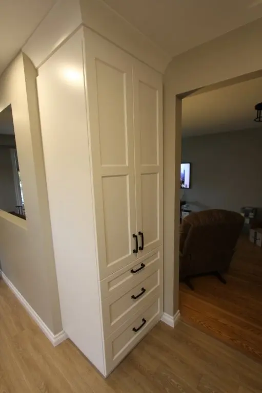 Tall white cabinet with double doors and three drawers featuring black handles, positioned in a corner between a hallway with light wood flooring and a living room with a brown armchair and TV.