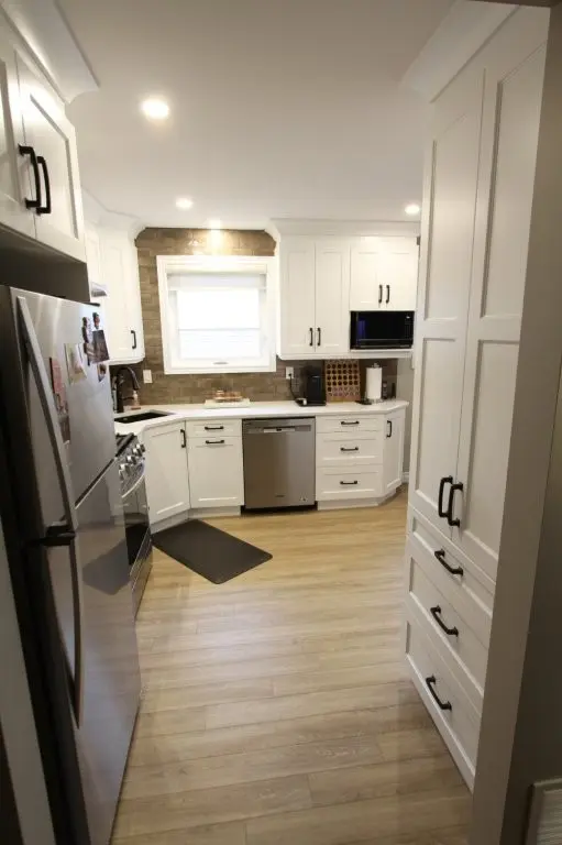 Modern kitchen with white cabinets, stainless steel appliances including a refrigerator, stove, and dishwasher, wooden flooring, under-cabinet lighting, and a window above the sink with a roller shade.