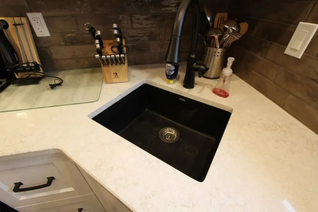 Black kitchen sink with a matte black faucet set in a beige countertop, accompanied by a wooden knife block, stainless steel utensil holder, and a soap dispenser with red liquid, against a brown tiled backsplash.