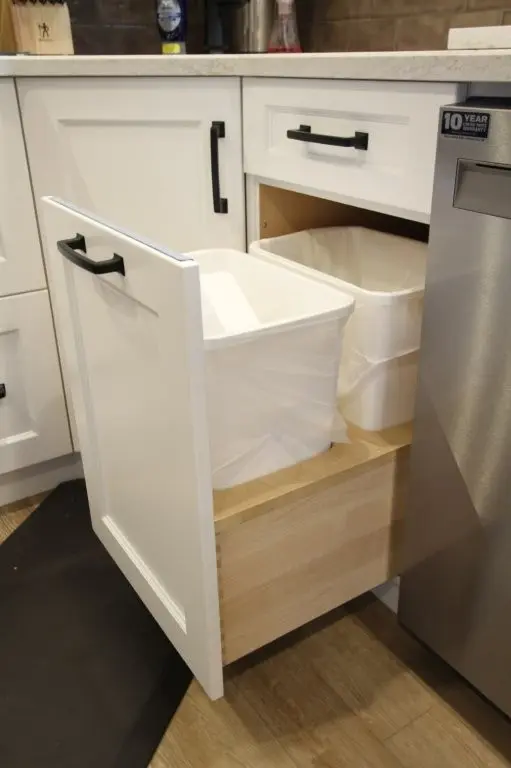 Pull-out kitchen cabinet with two white trash bins inside, one bin partially pulled out, white cabinetry with black handles and a stainless steel appliance next to it.