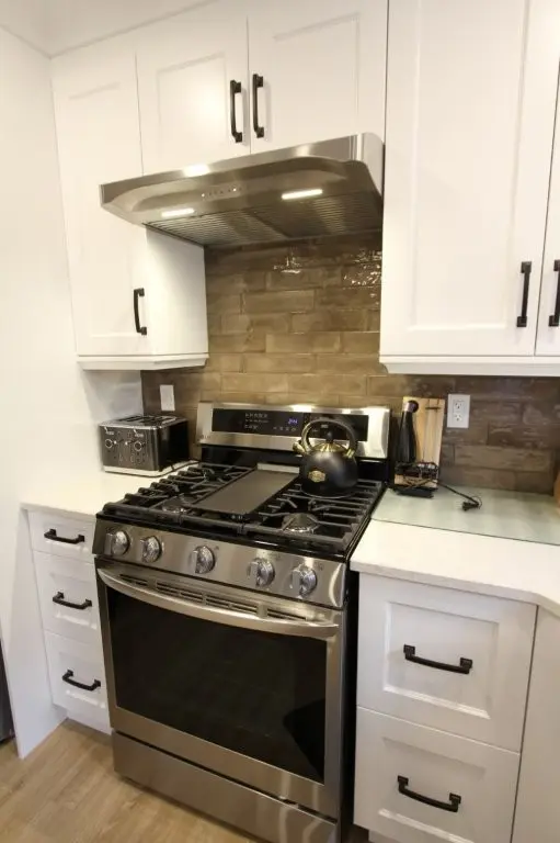 Modern kitchen with a stainless steel gas stove featuring four burners and a griddle, a black kettle on the front right burner, white cabinets with black handles, a stainless steel range hood with lights, a toaster, and brown subway tile backsplash.
