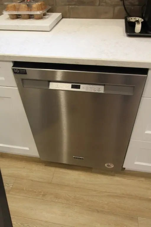 Stainless steel dishwasher installed under white countertop in a kitchen, flanked by white cabinets and placed on wood flooring.