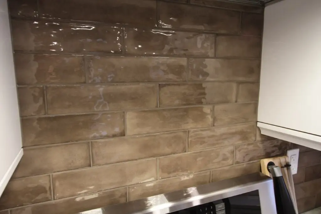 Glossy brown rectangular subway tiles backsplash in a kitchen corner above a stainless steel microwave, with white cabinets on each side.