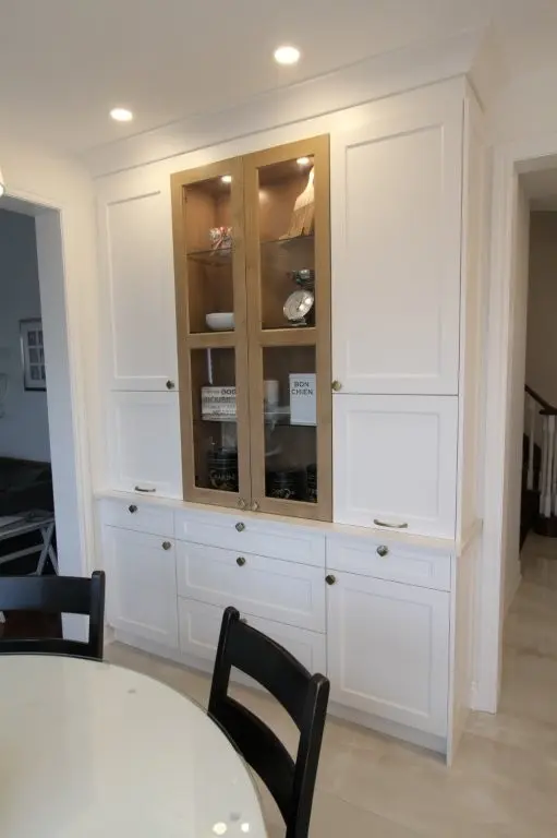 Built-in white kitchen cabinet with a central section featuring wooden-framed glass doors displaying dishes and decor items, next to a round glass-top table with black chairs in a well-lit room.