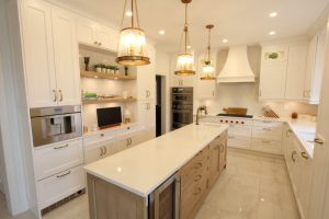 Bright modern kitchen with white cabinetry, a long central island with light wood base and white countertop, three pendant lights hanging above, built-in stainless steel appliances, and decorative plants on open shelves.