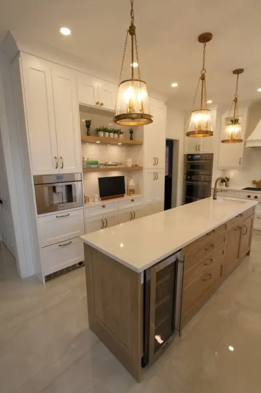 Modern kitchen interior with white and wood cabinetry, a large island with a white countertop and built-in wine fridge, three hanging pendant lights with warm bulbs, and stainless steel appliances.