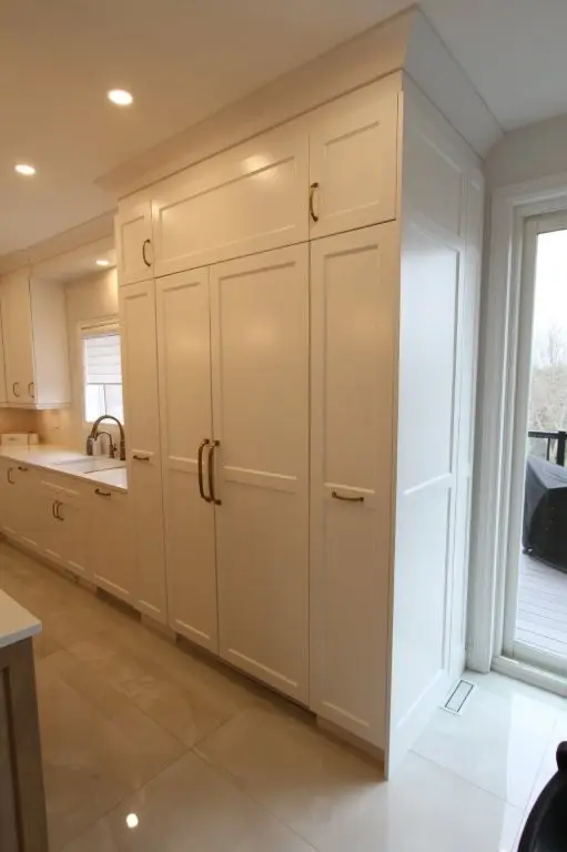 Tall white kitchen cabinets with gold handles, a built in fridge, a sink with a silver faucet under a window, and a sliding glass door leading to a balcony with a grill.