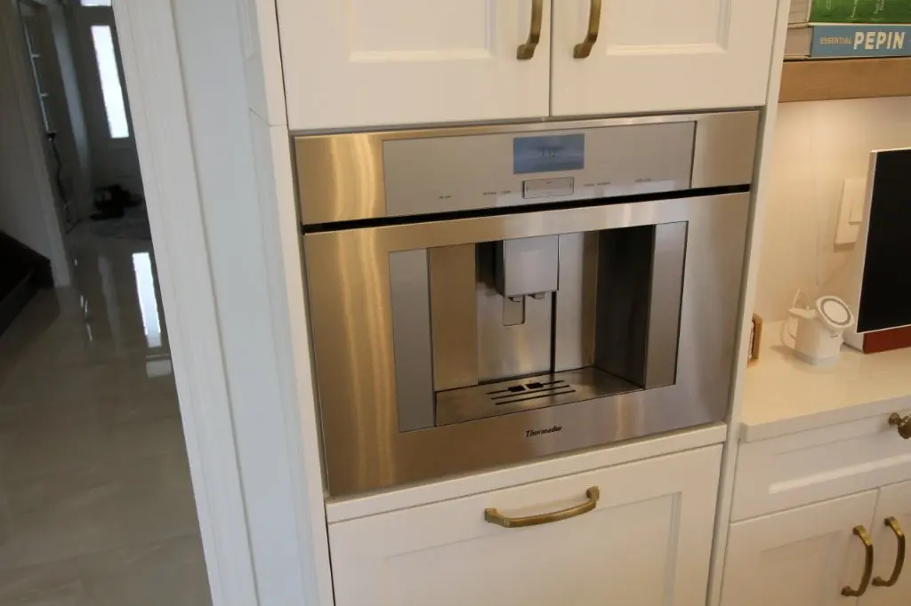 Built-in stainless steel Thermador coffee machine integrated into white kitchen cabinetry with gold handles, adjacent to a white countertop and a nearby monitor screen.