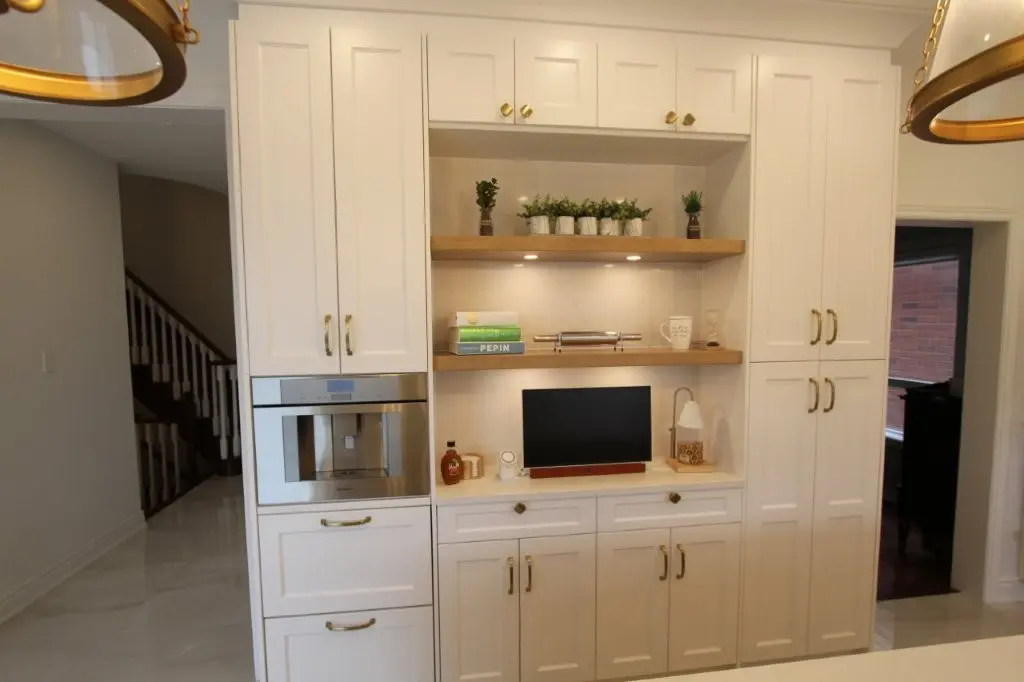 White built-in kitchen cabinets with gold handles, open shelves with small potted plants and books, a built-in coffee machine, a countertop with a small screen device, a lamp, and decorative items under warm lighting.