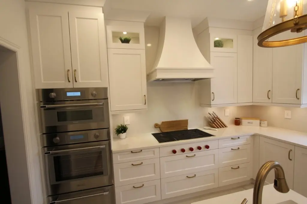 Modern white kitchen featuring a built-in stainless steel double oven, white cabinetry with brass handles, a gas cooktop with red knobs and a white vent hood above, and white countertops with decorative plants and kitchen items.