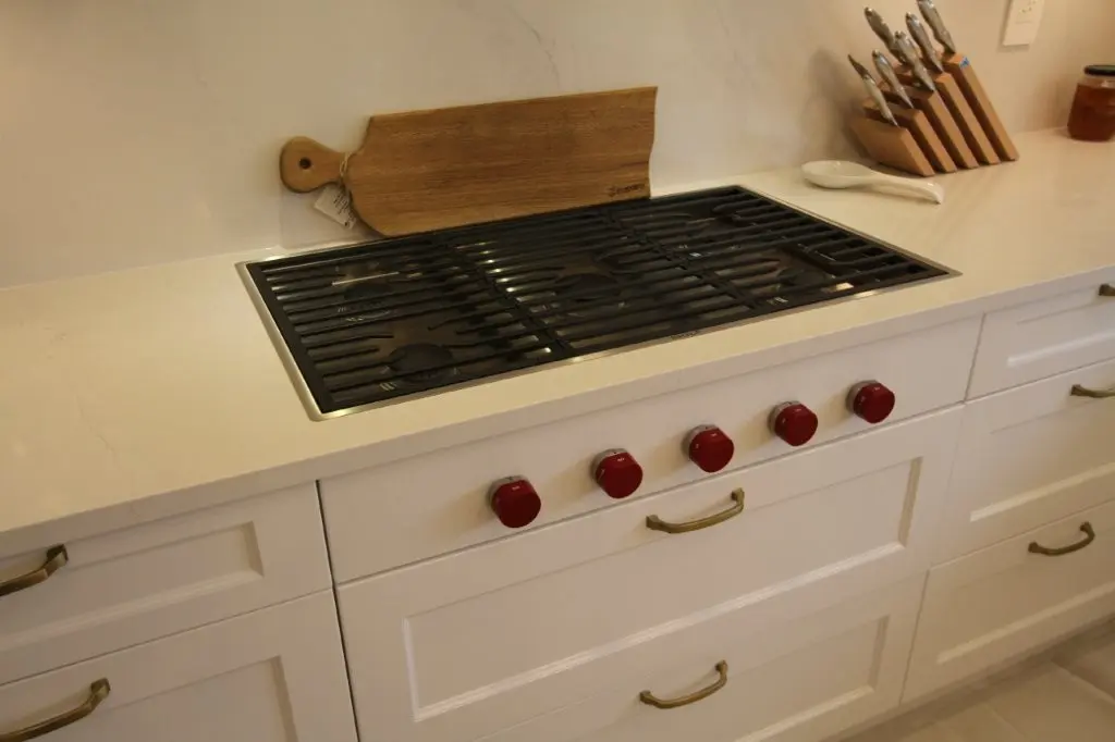 Modern kitchen countertop with a stainless steel gas stovetop featuring five red control knobs, white cabinetry with brass handles, a wooden cutting board leaning against the backsplash, and a knife block with knives in the background.