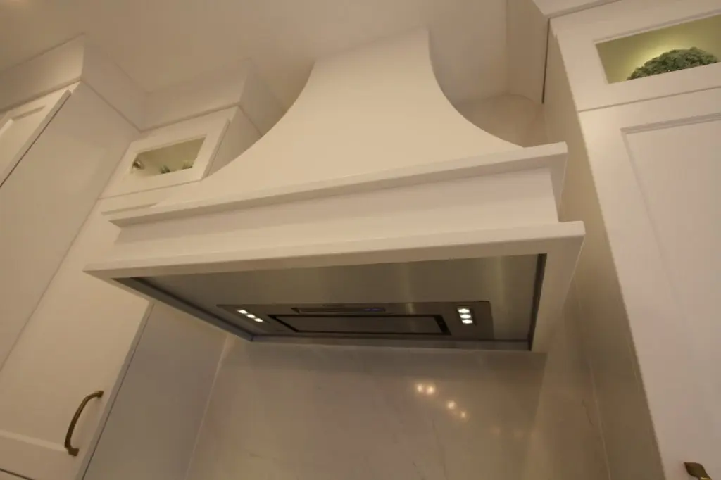 White kitchen range hood with built-in stainless steel vent and lights, surrounded by white cabinets with glass display sections.