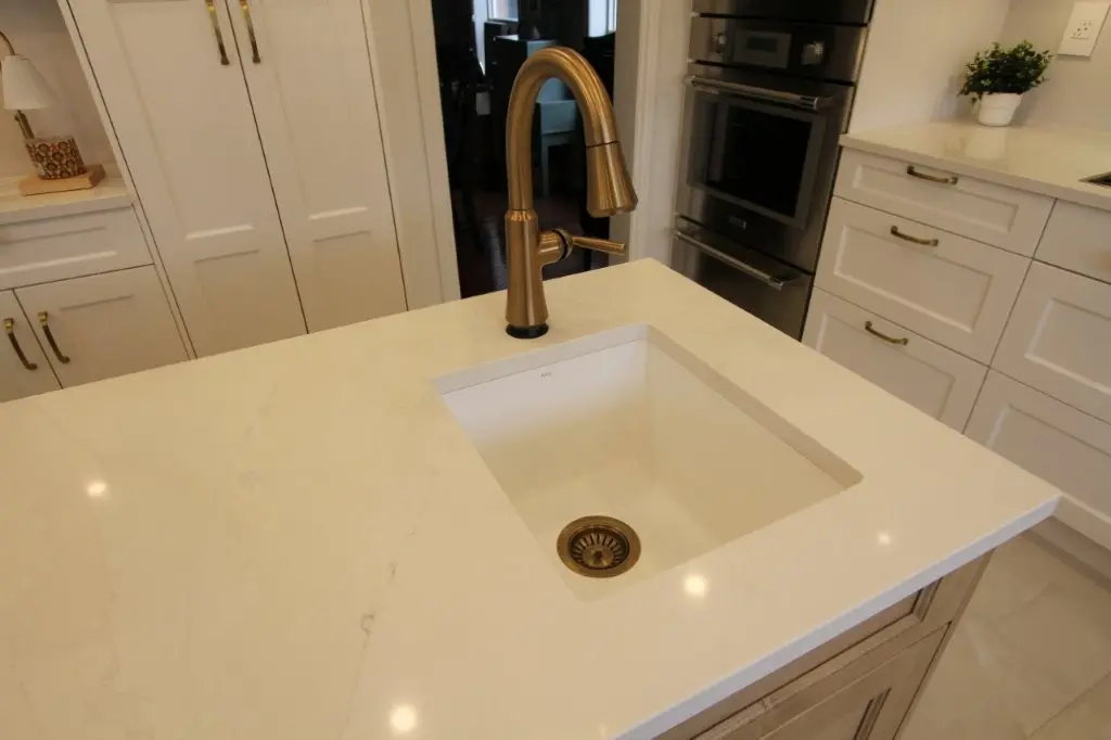 Modern kitchen sink with a matte gold faucet on a white marble countertop island, surrounded by white cabinetry and stainless steel built-in ovens in the background.