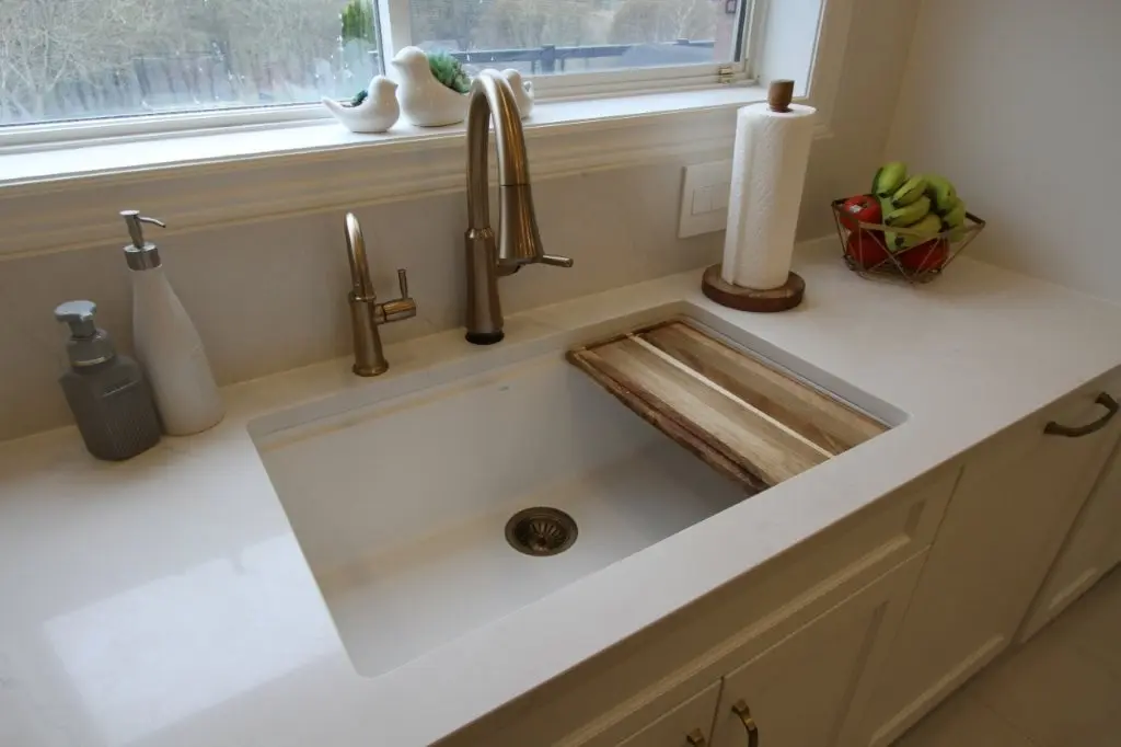 Modern kitchen sink with gold faucet, soap dispensers on the left, a wooden cutting board resting across the sink, a paper towel roll on the right, and a fruit basket with bananas and tomatoes on the countertop near the window.
