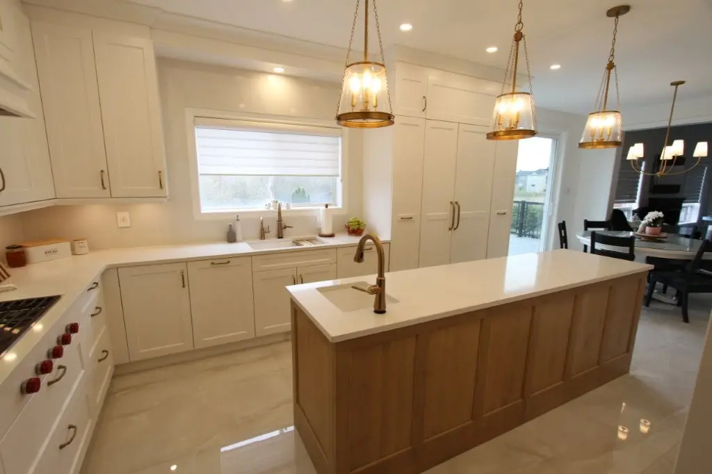 Modern kitchen with a large wooden island topped with a white countertop and a built-in sink, three pendant lights hanging above, white cabinetry along the walls, a window with a rolled-up blind above the sink, and a dining area with a chandelier and black chairs visible in the background.
