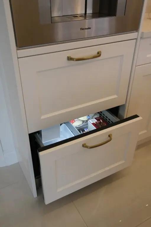 Modern kitchen with built-in stainless steel Thermador appliance above two white refrigerator drawers, the bottom drawer partially open revealing icemaker inside.