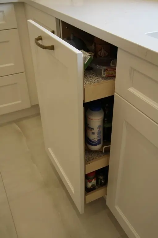 Partially open kitchen cabinet door revealing pull-out shelves stocked with various containers and kitchen items in a white cabinetry setting with a light countertop and tiled floor.