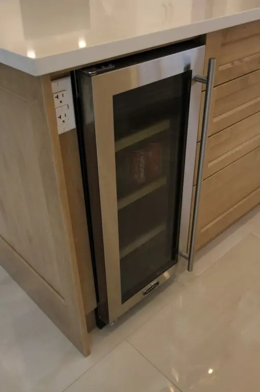 Built-in stainless steel beverage cooler with a glass door and metal handle integrated into wooden kitchen cabinetry, showing some cans inside.