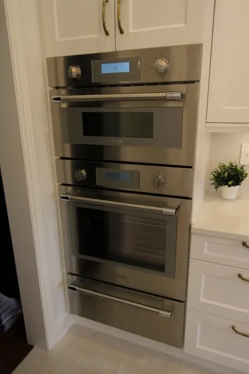 Stacked stainless steel built-in kitchen ovens integrated into white cabinetry with digital and dial controls, next to a white countertop holding a small potted plant.