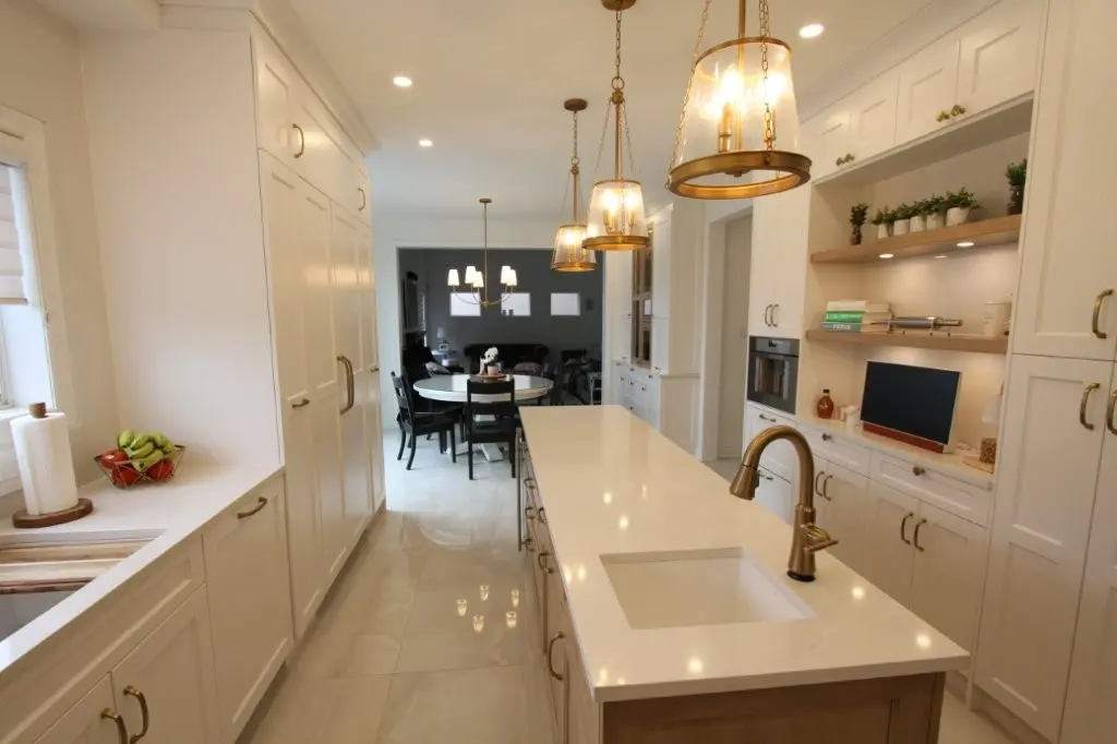 Bright modern kitchen with white cabinetry, a large central island with a built-in sink and brass faucet, pendant lights hanging above, and a dining area with a round table and black chairs visible in the background.