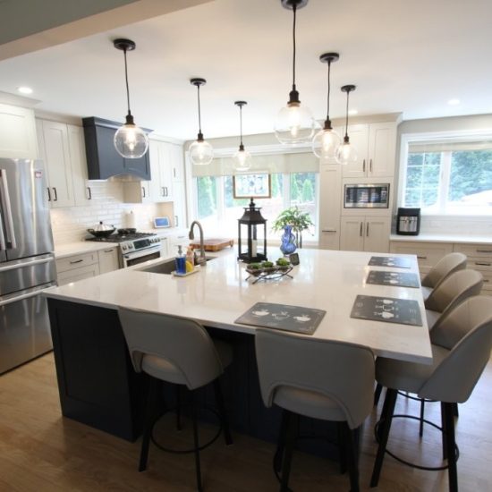 Modern kitchen with a large white island countertop surrounded by six beige bar stools, stainless steel refrigerator, white cabinets, hanging globe pendant lights, and large windows letting in natural light.