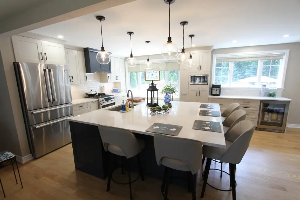 Modern kitchen with a large white island countertop surrounded by six beige bar stools, stainless steel refrigerator, white cabinets, hanging globe pendant lights, and large windows letting in natural light.