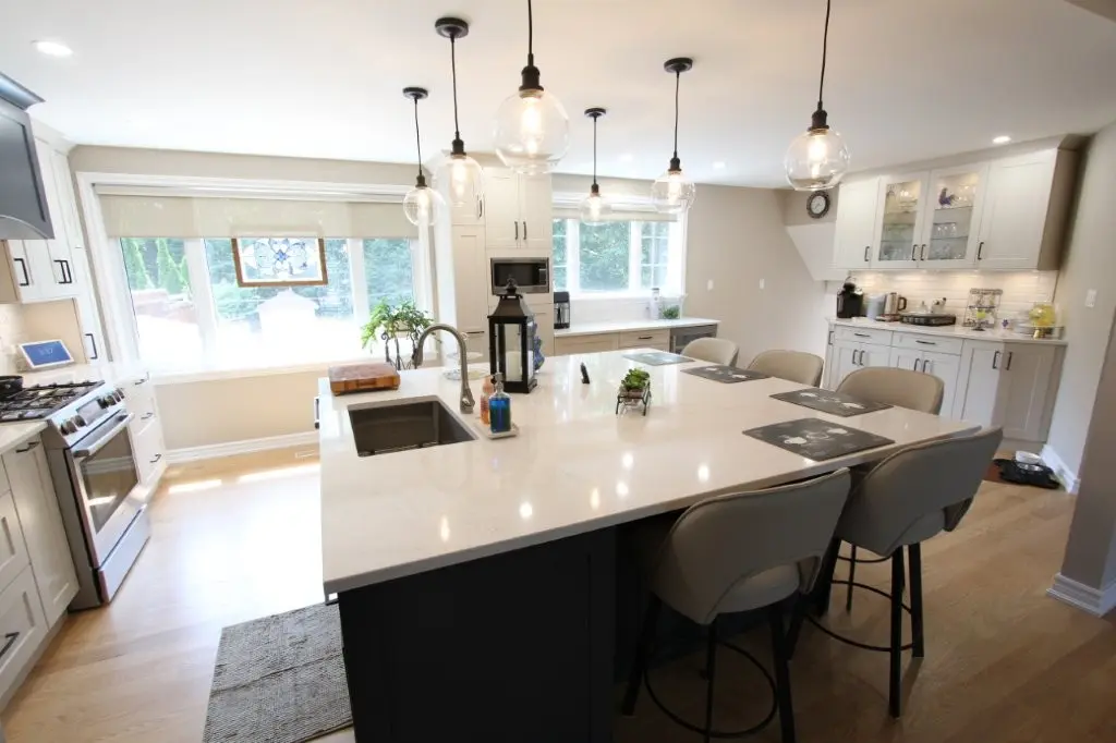 A bright modern kitchen featuring a large white island with a built-in sink, pendant lights hanging above, four beige bar stools, stainless steel appliances, light wood flooring, and large windows letting in natural light.
