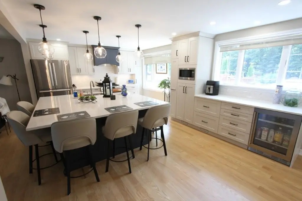 Bright modern kitchen with white cabinetry, a large island with six beige stools, pendant lights hanging above, stainless steel refrigerator, built-in microwave, and a window overlooking greenery.