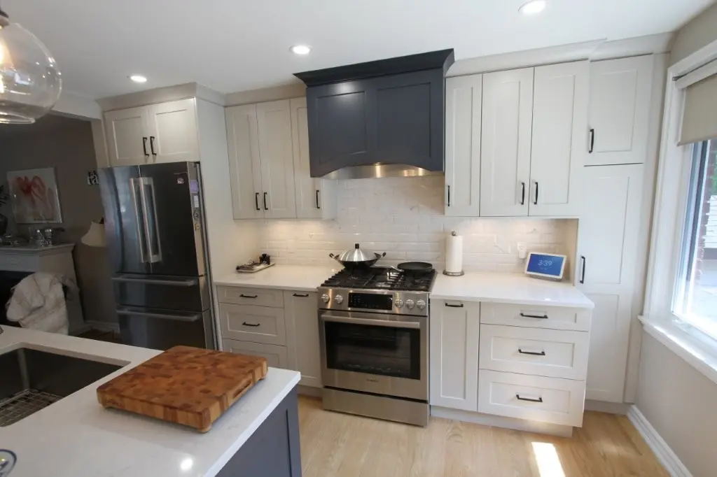 Modern kitchen with light gray cabinets, a stainless steel stove with a black range hood, a large refrigerator, and a wooden cutting board on a white countertop island near a sink, with a window on the right.