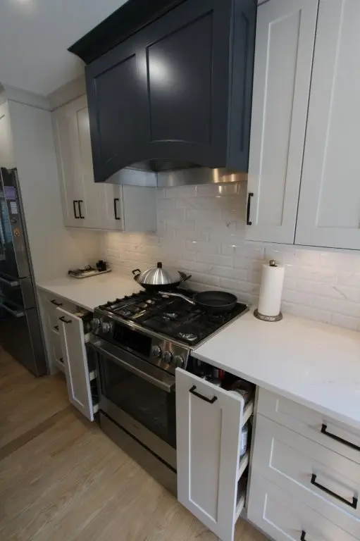 Modern kitchen stove area with a stainless steel gas range, black pan and pot on burners, white countertop, white tiled backsplash, white cabinetry with black handles, open cabinet doors showing spices, and a black range hood above.