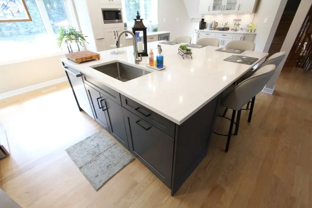 Modern kitchen island with white marble countertop, built-in sink with faucet, soap dispensers, a small plant, and gray cabinetry, surrounded by beige cushioned bar stools on light wood flooring.
