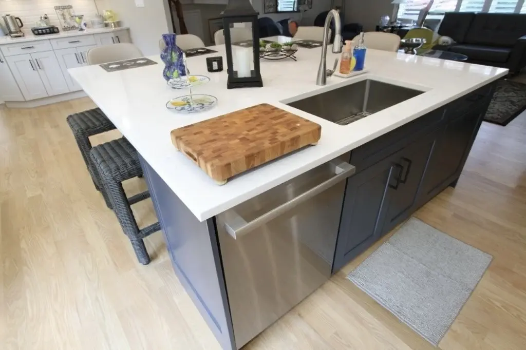 Modern kitchen island with a white countertop, a wooden cutting board, a stainless steel sink with a faucet, a lantern, a blue vase, hand soaps, and two wicker bar stools against light wood flooring