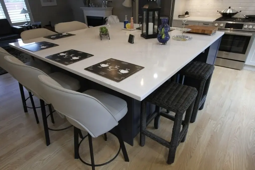 Modern kitchen island with a white marble countertop surrounded by four white cushioned chairs and two black wicker stools, set with four black placemats featuring white teapot designs, and decorated with a small potted plant, a black lantern, a blue glass vase, and kitchen items in the background including a stove and a wooden cutting board.