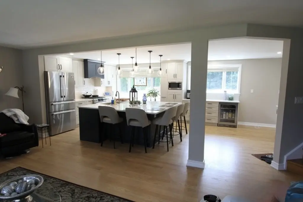Modern kitchen with white cabinetry, a large island with bar stools, stainless steel refrigerator, wood flooring, and pendant lights hanging over the island, viewed from an adjacent living area.