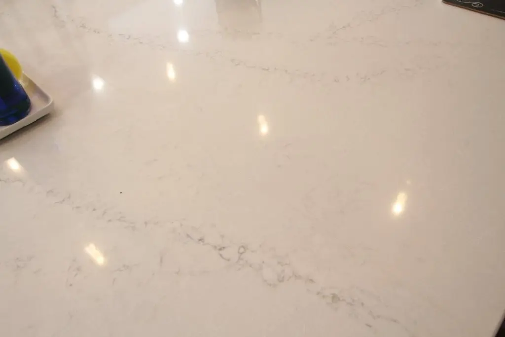 Smooth white marble countertop with subtle gray veins and reflections of overhead lights, with a partial view of a tray holding bottles on the left corner