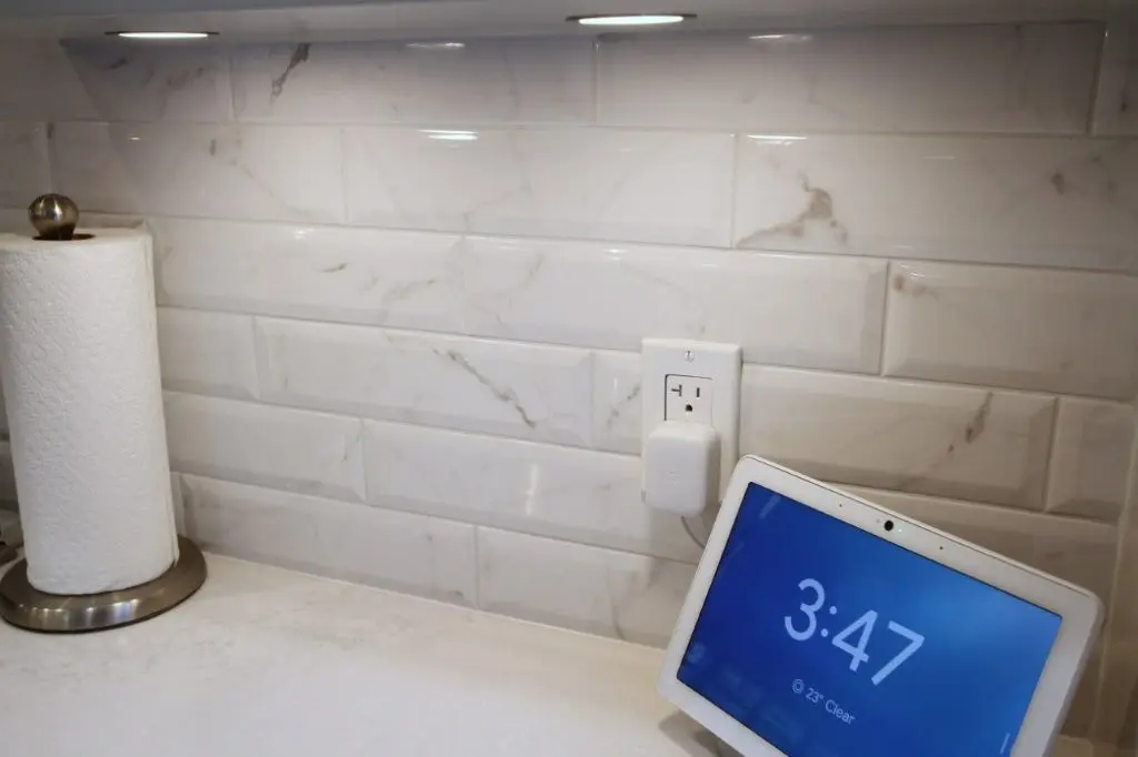 Smart display device showing time 3:47 and weather 23° Clear on a kitchen countertop next to a paper towel holder, with a marble-patterned tile backsplash and a plugged-in smart home device on the wall outlet.