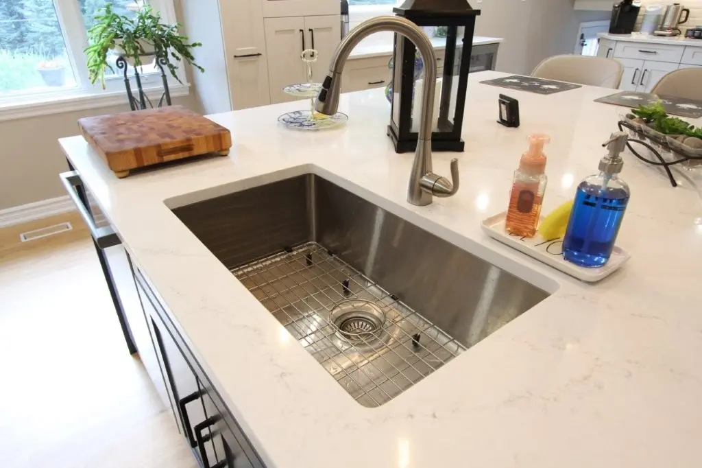Modern kitchen island with a white marble countertop featuring a built-in stainless steel sink and a curved faucet, a wooden cutting board to the left, and soap dispensers on a tray to the right, with a plant and window in the background.