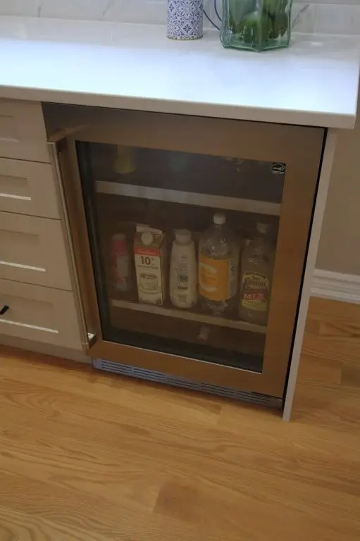 Built-in beverage cooler with a glass door in a kitchen cabinet, containing various bottles and cartons on wooden shelves, with a white countertop above and light wood flooring below.