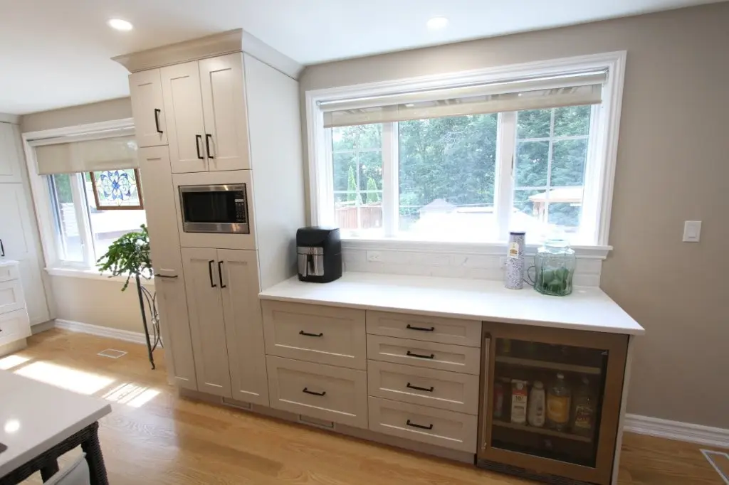 Modern kitchen cabinetry with built-in microwave, air fryer, storage drawers, and a glass-door beverage cooler under a large window with a view of trees outside.