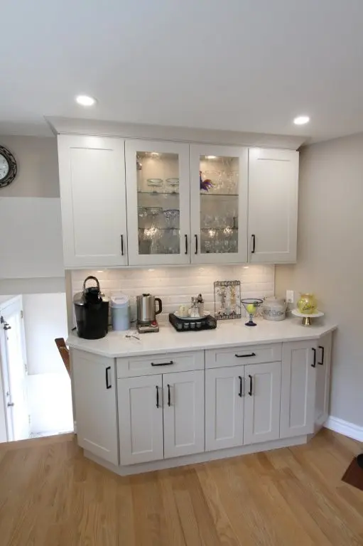 White kitchen cabinet with black handles featuring glass doors displaying glassware, a coffee machine, kettle, and decorative items on the countertop, set against a beige wall with wooden flooring.