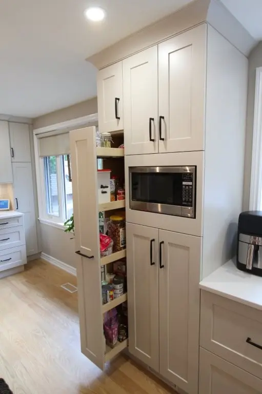 Kitchen pantry cabinet with beige doors, one door open showing organized shelves of spices and canned goods, built-in microwave above, and light wood flooring with window in the background.