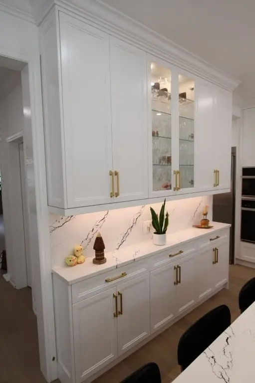 White kitchen cabinetry with gold handles and glass doors displaying glassware, a white marble countertop and backsplash with black veins, decorated with a potted snake plant and small figurines, set against a wooden floor.