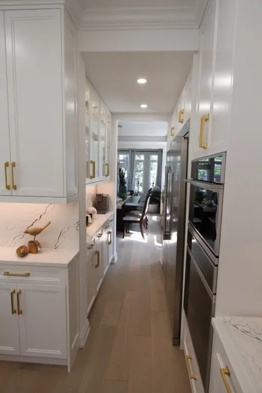 Narrow modern kitchen with white cabinetry featuring gold handles, marble-patterned countertops, built-in stainless steel oven, and light wood flooring leading to a dining area with chairs and glass doors allowing natural light.