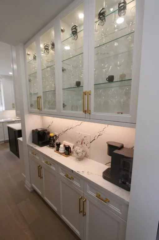 A kitchen nook with white cabinets featuring gold handles, glass-front upper cabinets displaying glassware, a white marble countertop with black veining holding a coffee machine, a coffee pod holder, and other small items, all under soft recessed lighting.