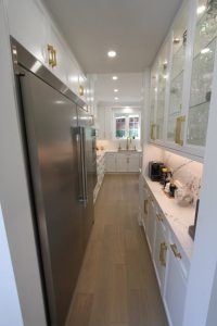 Narrow modern kitchen with light wood flooring, white cabinetry with gold handles, a large stainless steel refrigerator on the left, glass-front upper cabinets displaying glassware on the right, and a white marble countertop with a coffee machine and decorative items, with a window at the far end.