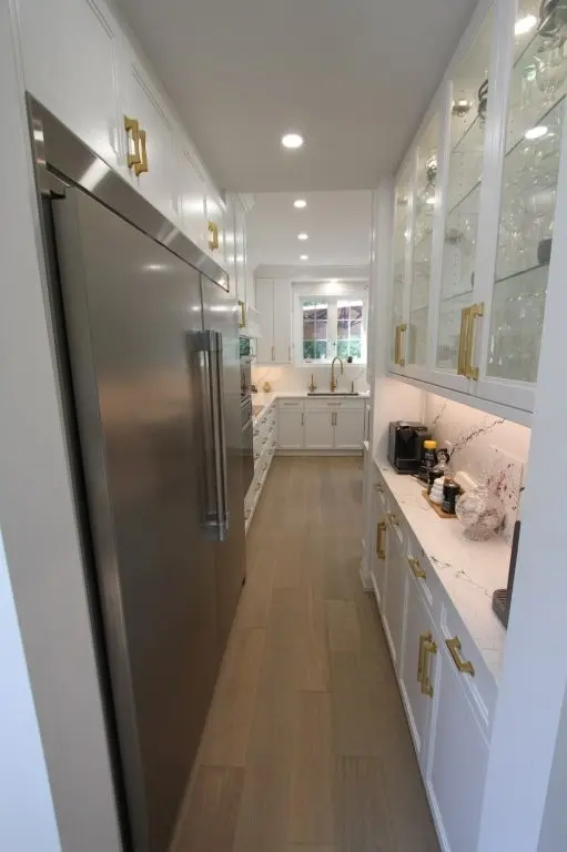 Narrow modern kitchen with light wood flooring, white cabinetry with gold handles, a large stainless steel refrigerator on the left, glass-front upper cabinets displaying glassware on the right, and a white marble countertop with a coffee machine and decorative items, with a window at the far end.