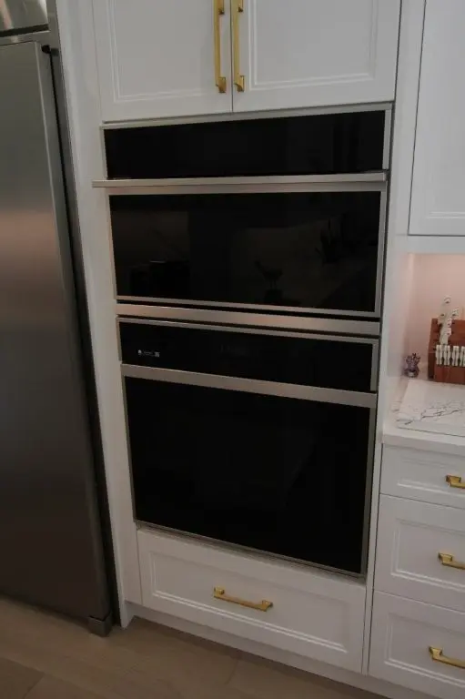 Built-in modern kitchen appliances including a black and stainless steel oven and microwave fitted within white cabinetry with gold handles, next to a stainless steel refrigerator and white marble countertop with a wooden knife block and utensils on the side.